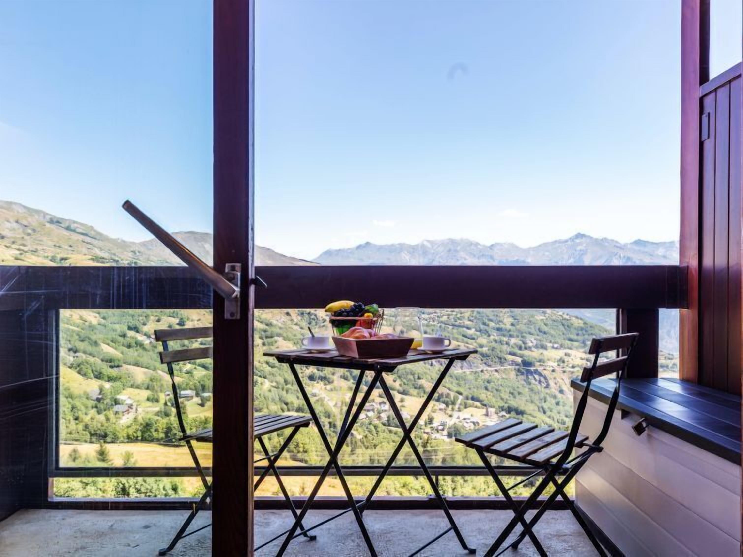 Een balkon met een tafel en stoelen, een schaal fruit en uitzicht op een bergachtig landschap.