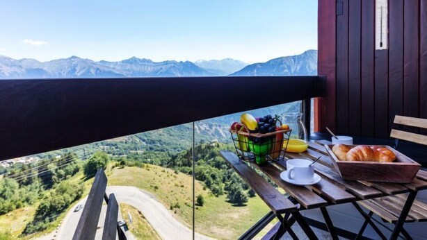 Un balcon avec vue sur les montagnes, une table avec un panier de fruits et une miche de pain.