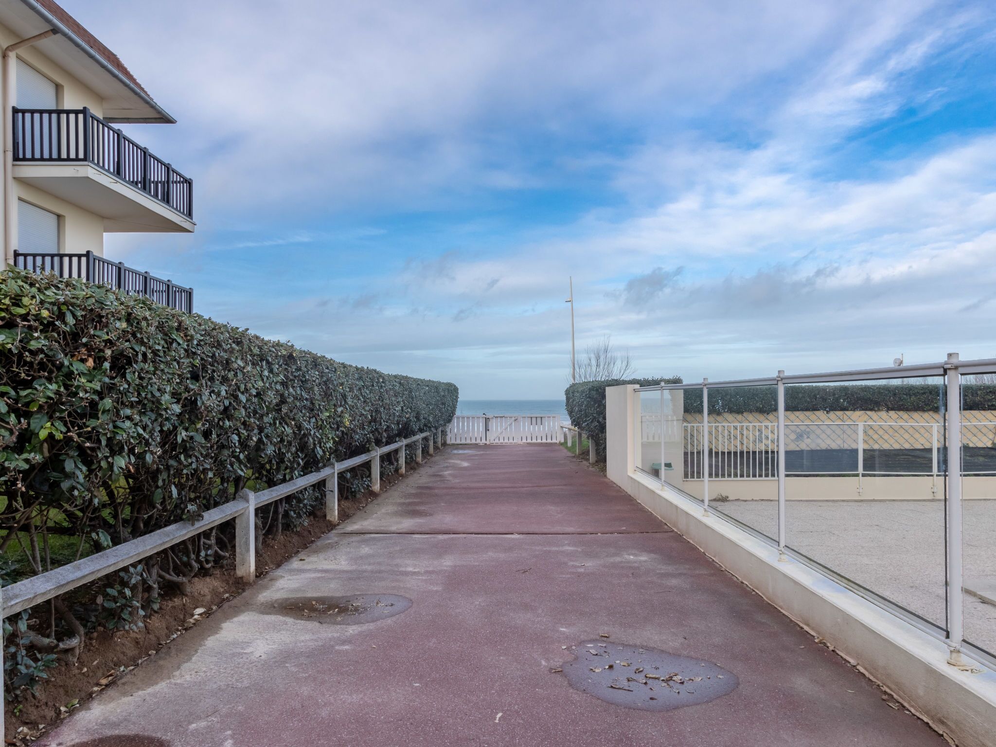 A pathway leading to a beach with a fence on one side and a building with a balcony on the other.