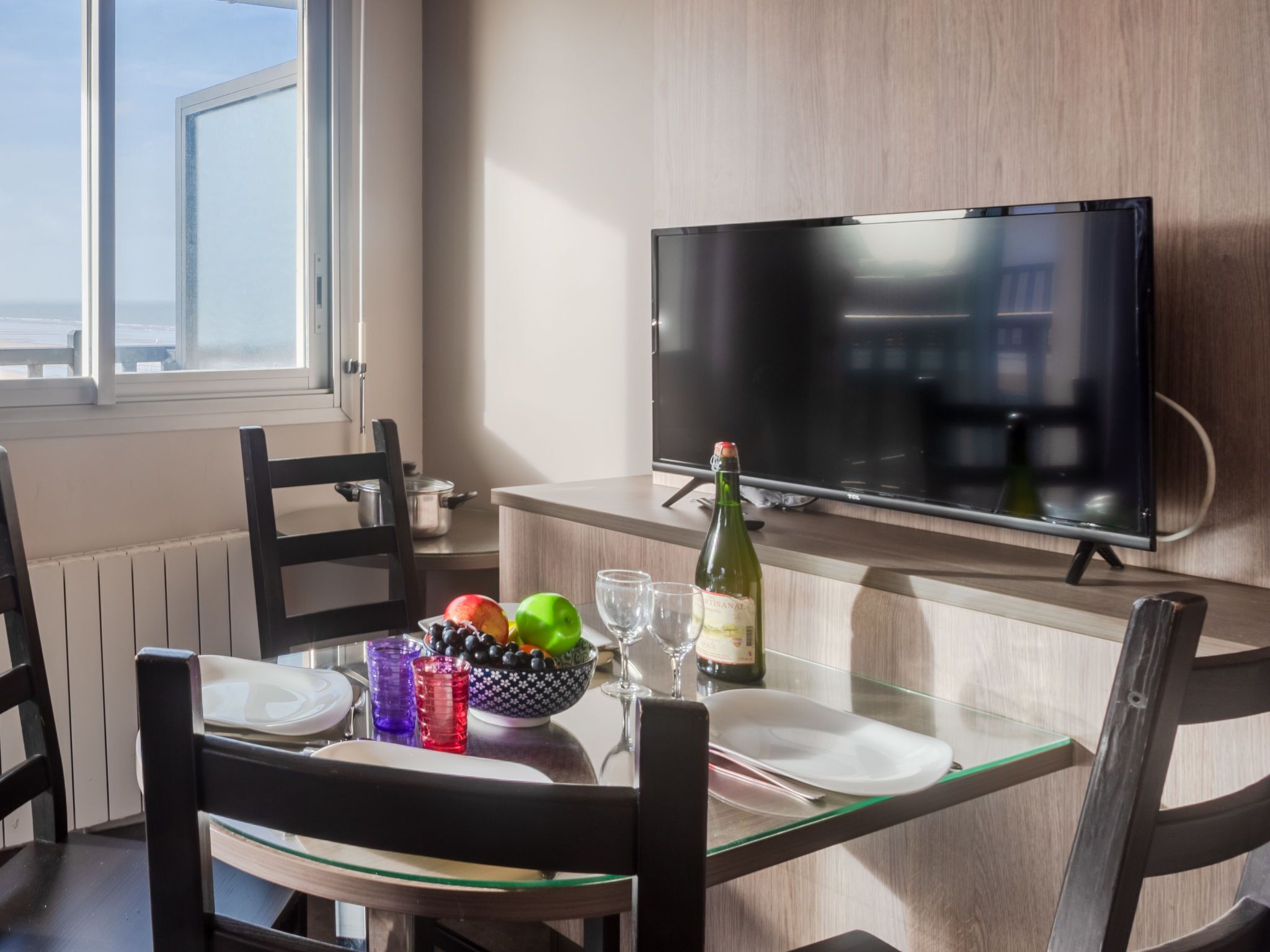 A dining area with a table set for two, a bottle of wine, a bowl of fruit, and a television on the wall.