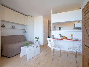 A compact living space with a sofa, a small table with a plant, a dining area with chairs, and a kitchenette with a microwave and a small fridge.