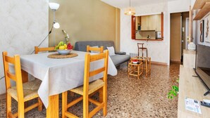 Un espace repas comprenant une table, des chaises, un saladier de fruits, un canapé, un coin cuisine avec un miroir et un téléviseur.