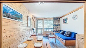 Ein gemütliches Blockhaus-Interieur mit Holzwänden, einer Essecke mit Tisch für vier Personen, einem blauen Sofa und einem großen Fenster mit Blick ins Freie.