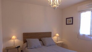 Un dormitorio con una cama individual, dos mesitas de noche, una araña, un cuadro en la pared y una ventana con cortinas.