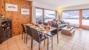 En matplats med en väggpanel av trä, ett bord dukat med tallrikar, koppar och en korg med frukt. Det finns en soffa med kuddar och en utsikt över bergen genom stora fönster.