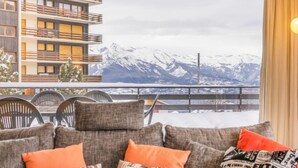 En balkong med utsikt över snöklädda berg, en soffa med mönstrade kuddar och en matplats med stolar.