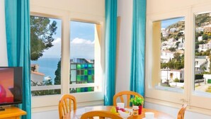 A dining area with a table and chairs, a television on a stand, and a view of a coastal town through large windows. - Velazquez by Interhome (Roses)