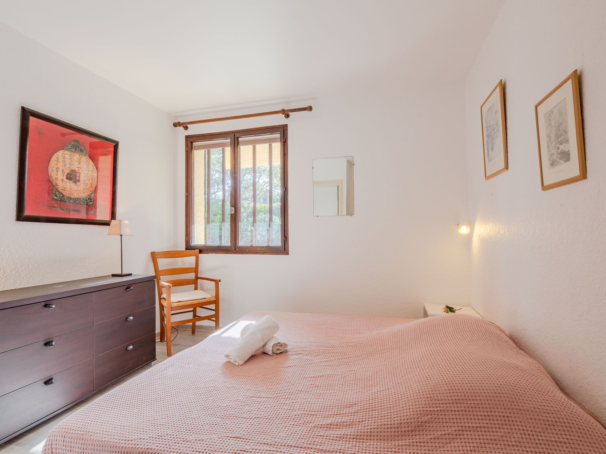A bedroom with a bed, a wooden chair, a dresser, a window, and wall-mounted artwork.