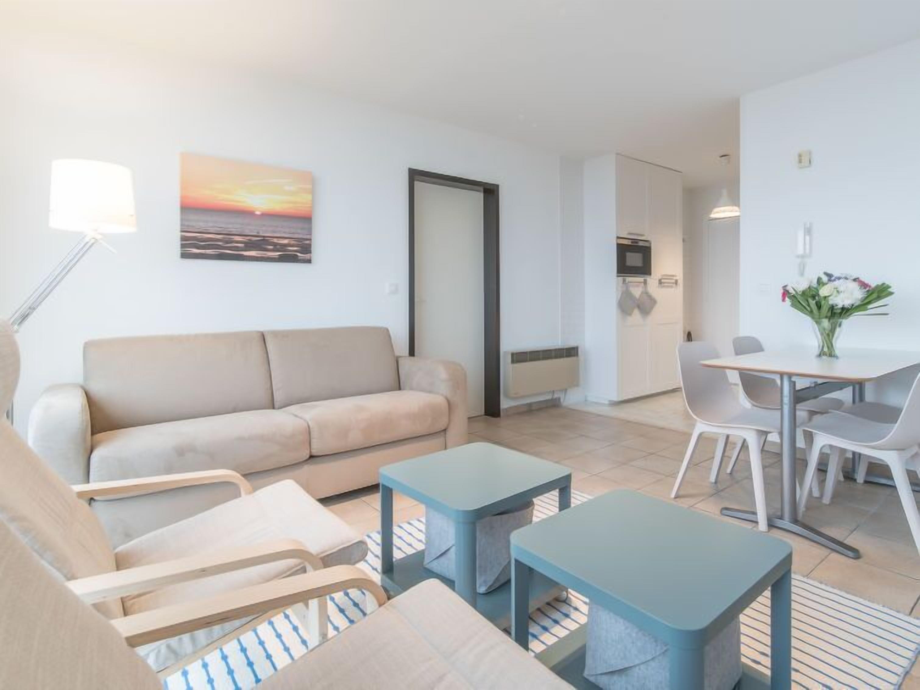 Uma sala de estar moderna com um sofá bege, uma mesa de jantar com cadeiras e uma pequena cozinha com televisão e micro-ondas.