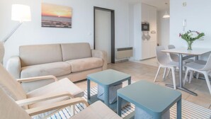 Una sala de estar moderna con un sofá color beige, una mesa de comedor con sillas y una pequeña cocina con televisor y microondas.