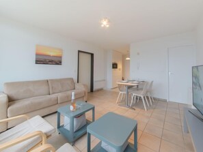 A modern living room with a beige sofa, a small table with a bottle, a TV, and a dining area with chairs.