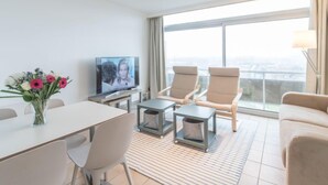 Habitación de hotel moderna con comedor, televisor, dos sillones, un sofá y un ventanal con vistas a la ciudad.