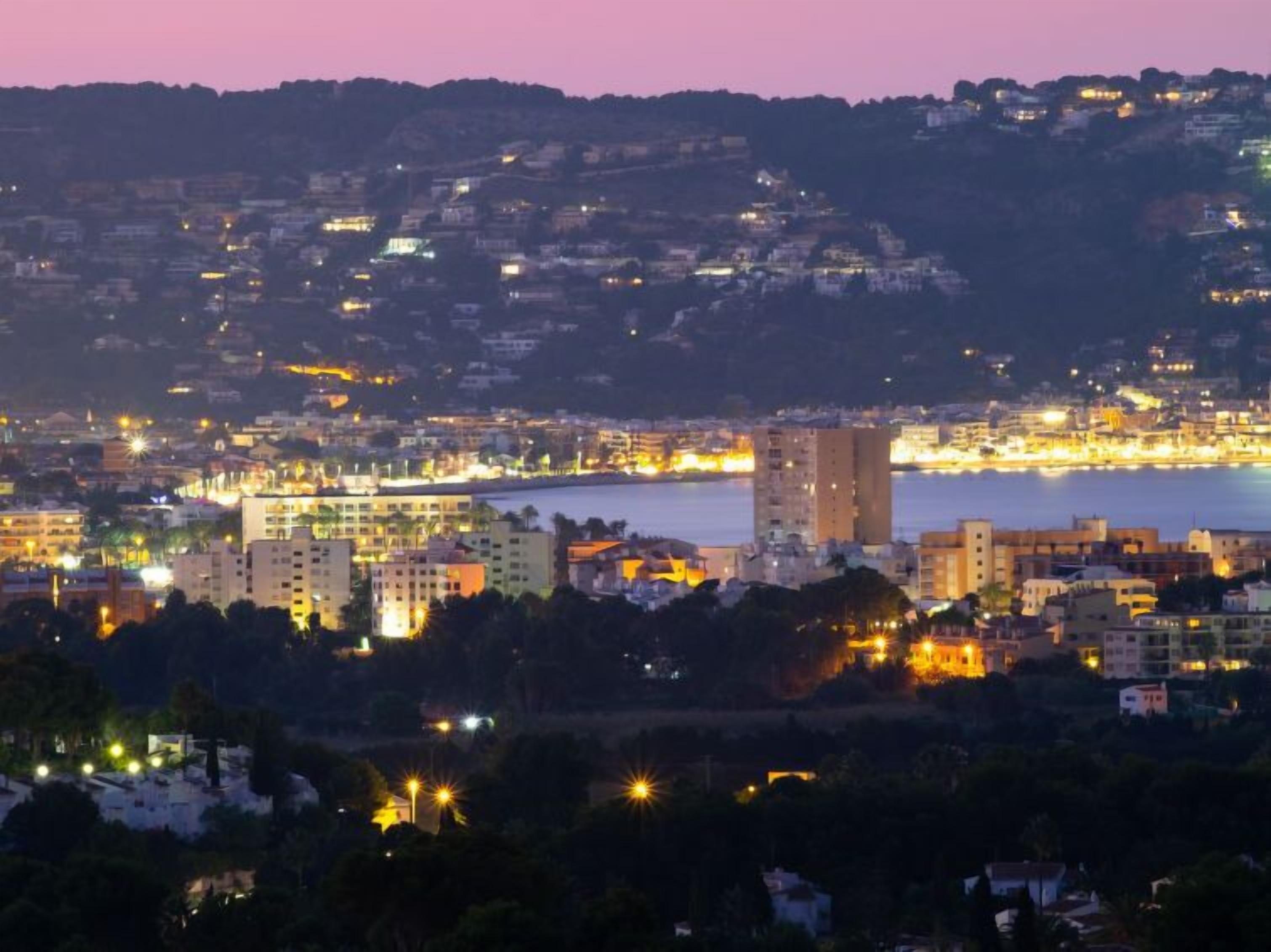 A coastal cityscape at dusk with buildings along the waterfront and a hilly backdrop.