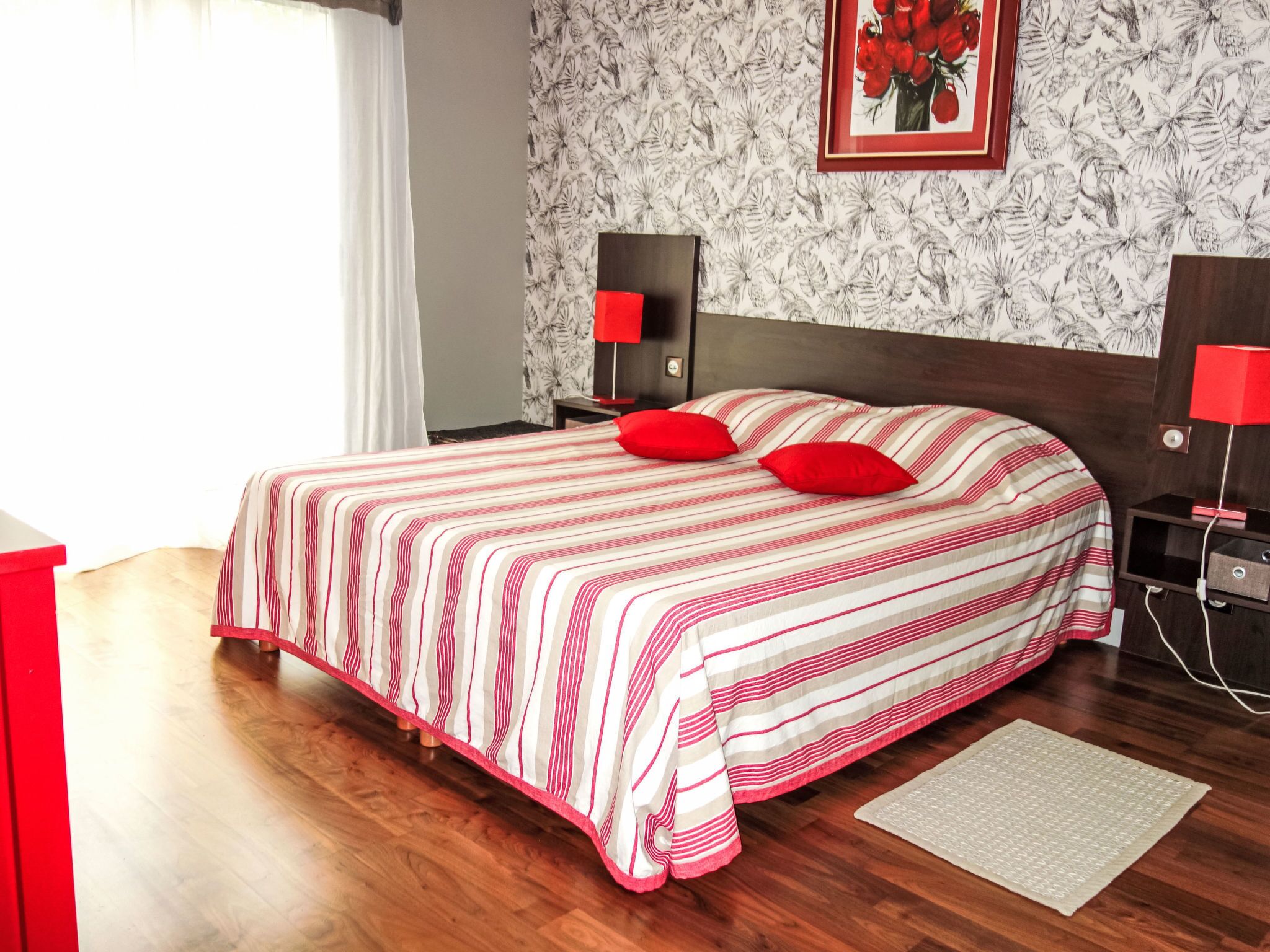 Une chambre d’hôtel avec un grand lit, deux tables de chevet, une commode, une lampe rouge, un tableau encadré au mur et une fenêtre avec des rideaux.