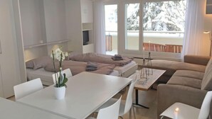 A modern, well-lit room with a dining table, chairs, a sofa, and a TV. Large windows provide natural light and a view of the outdoors. - Val Signina by Interhome (Laax)