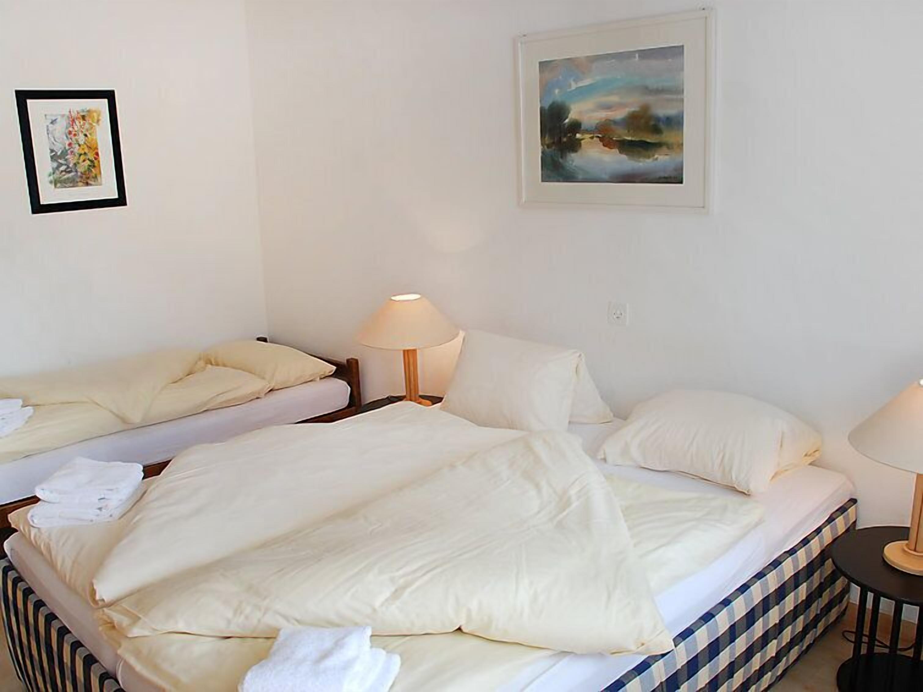 Ein Hotelzimmer mit zwei Betten, weißen Leinentüchern, einem Gemälde an der Wand und einer Lampe auf jeder Bettseite.