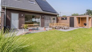 Une maison moderne avec un toit en pente, des panneaux solaires, une terrasse avec des meubles en bois et une petite structure en bois.