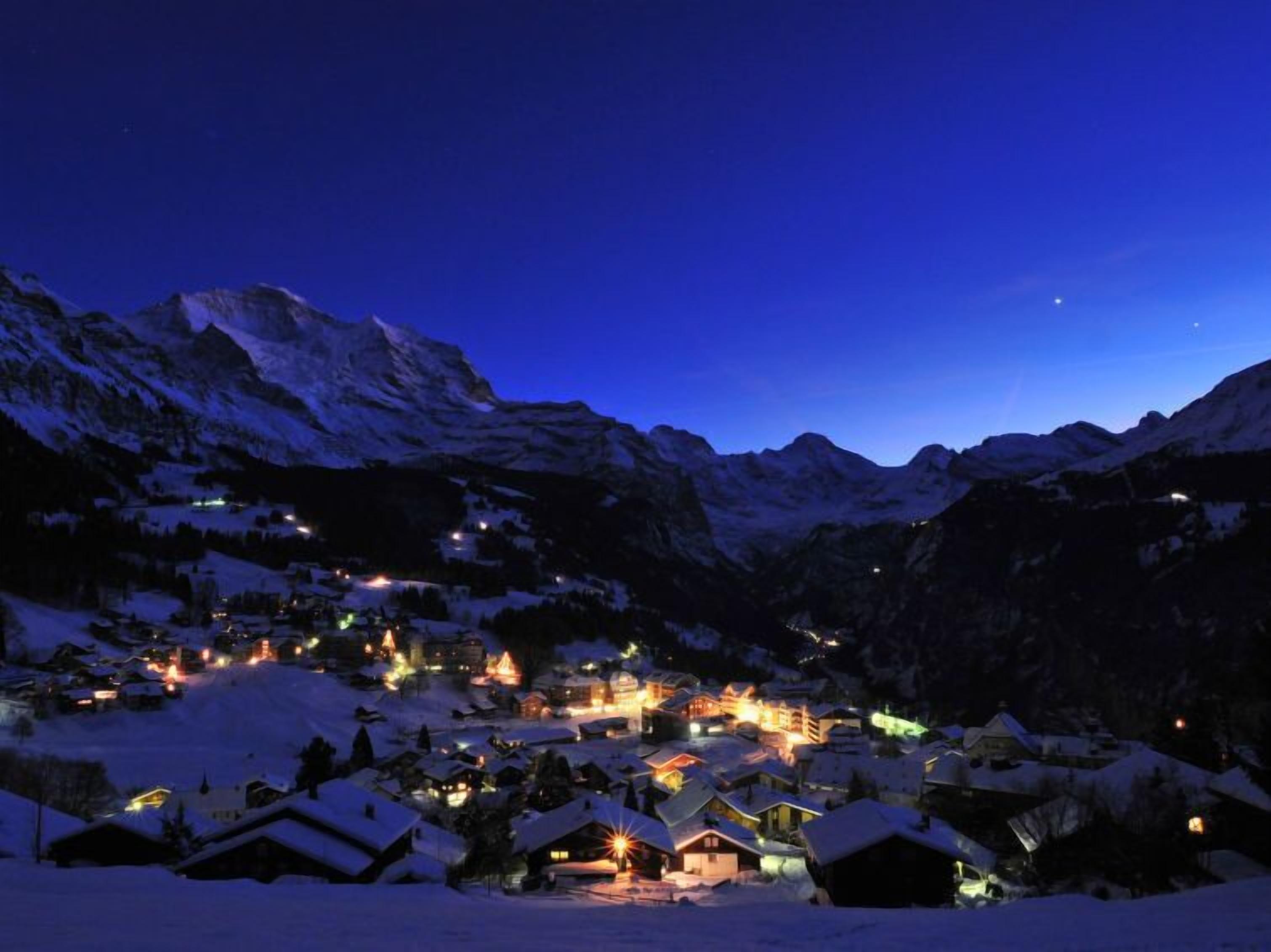 A snow-covered village at night with illuminated buildings and mountains in the background.