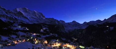 A snow-covered village at night with illuminated buildings and mountains in the background.