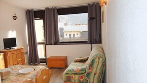 Een knus kamertje met een houten eettafel gedekt voor twee personen, een bank met een kleurrijk kussen, een televisie op een houten kast en een raam met gordijnen.