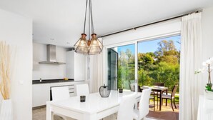 A modern kitchen with white cabinets, a dining area with a white table and chairs, and a large sliding glass door leading to an outdoor patio with a view of greenery. - Los Naranjos Singles by Interhome (Marbella)