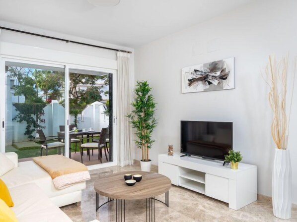 A modern living room with a sofa, a coffee table, a TV on a white stand, and a sliding glass door leading to an outdoor area with a table and chairs.