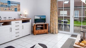 En moderne stue med fjernsyn, træunderholdningsmøbel, hvide skabe og en stor skydedør til en udendørs terrasse.