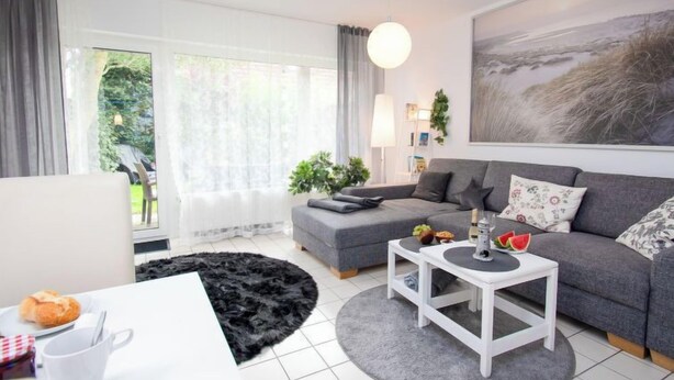 A modern living room with a grey sofa, a white coffee table, and a black rug. There is a window with sheer curtains, a painting on the wall, and a plant on the side table. - Muschelweg by Interhome (Norddeich)