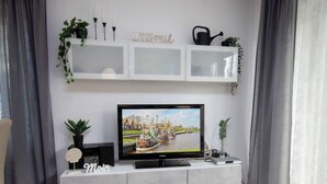 A modern living room with a flat-screen TV on a grey console, a plant, and a shelf with decorative items. - Muschelweg by Interhome (Norddeich)