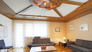 Un salon spacieux doté d’un plafond en bois, d’une grande fenêtre ornée de rideaux légers, de deux canapés, d’une table basse sur laquelle sont disposés des fruits, et d’une petite table avec une lampe.