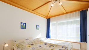 Une chambre avec un plafond en bois, un lit recouvert d’une literie à motifs floraux, une table de chevet avec une lampe, et deux tableaux accrochés au mur.