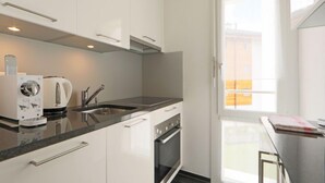 Une cuisine moderne avec des meubles blancs, un four encastré, une machine à café et une fenêtre donnant sur des immeubles.