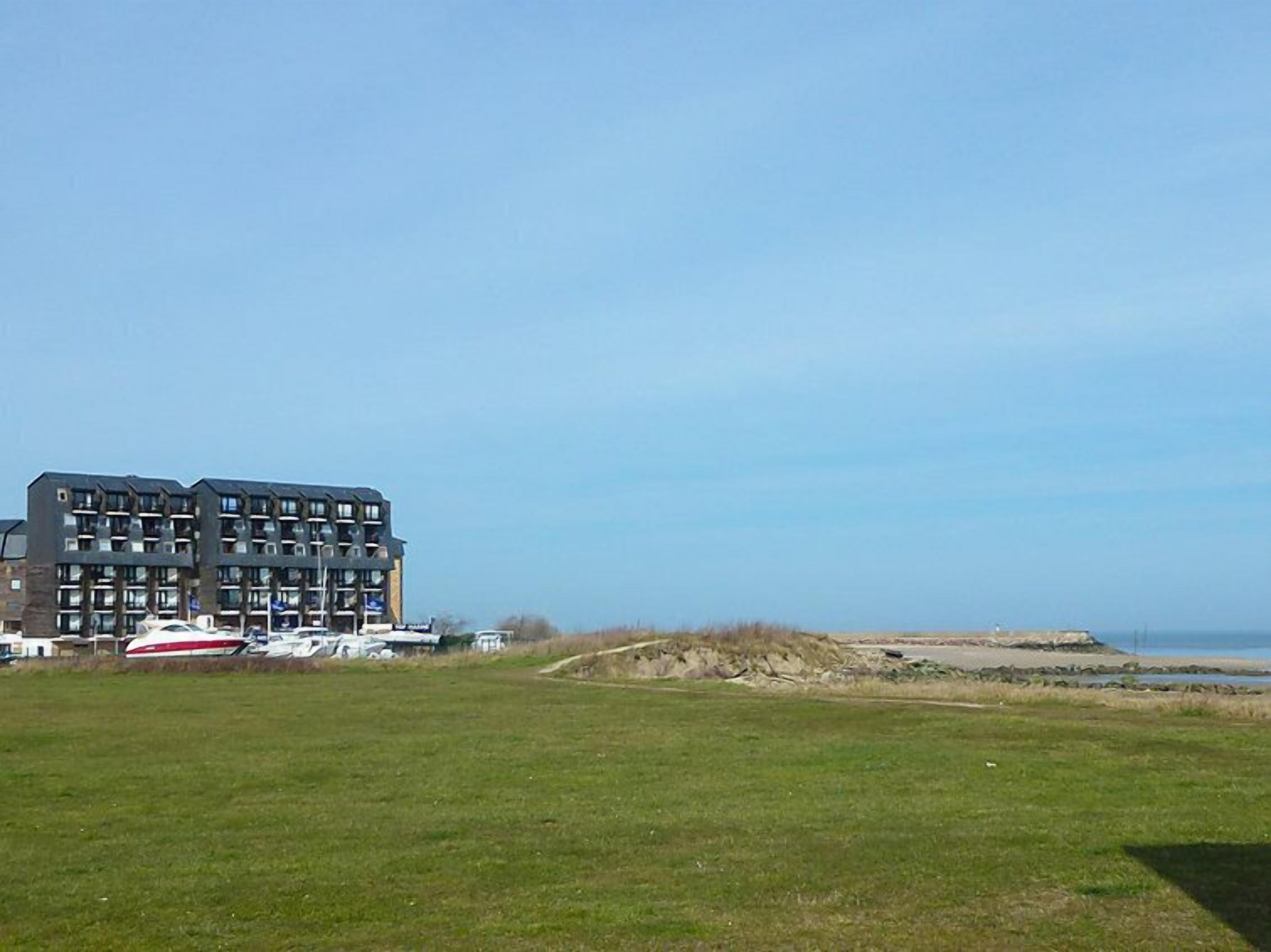 A modern multi-story building with a flat roof, located near a beach with a grassy field in the foreground.