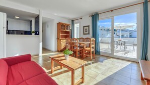 A modern living room with a red sofa, wooden dining table, and chairs. There is a kitchen area with cabinets and a window. The room has a sliding glass door leading to an outdoor patio with a table and chairs.