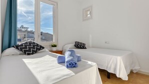 A room with two beds, a window with curtains, a small table with a plant, and a wall-mounted light fixture.
