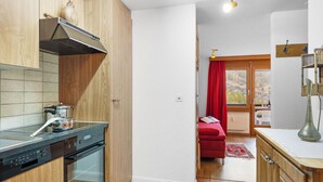 A compact kitchen with wooden cabinets, a built-in oven, and a stove. A red curtain and a small red sofa are visible in the adjacent living area.