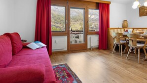A cozy living room with a red sofa, wooden dining table with chairs, and a view of mountains through large windows.