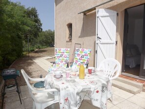 Une terrasse extérieure avec une table dressée pour deux personnes, entourée de chaises blanches, d’une nappe blanche et d’une bouteille d’eau.