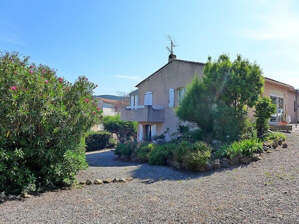A two-story house with a gravel driveway, surrounded by various shrubs and plants.