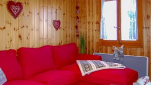 A red sofa in a room with wooden walls, a window with a view, and a small table with a star-shaped cushion. - Le Ruisseau by Interhome (Nendaz)