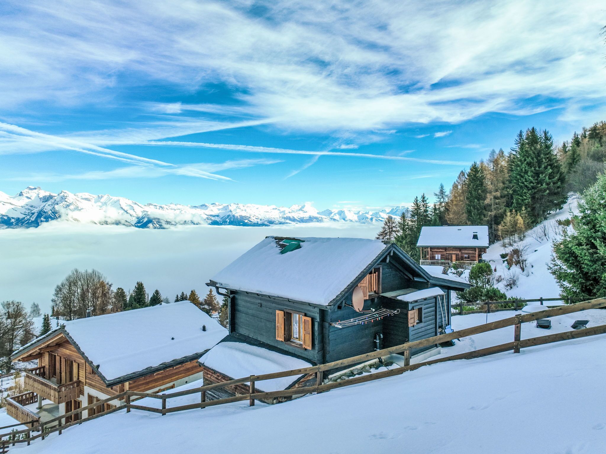 A wooden cabin with a balcony, surrounded by snow-covered trees and mountains in the background.