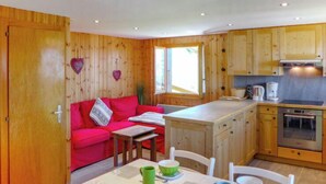 A cozy room with a red sofa, a small table with cups and a green mug, wooden cabinets, and a kitchen area with a stove. - Le Ruisseau by Interhome (Nendaz)