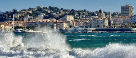 Un paysage urbain côtier avec des bâtiments, une église imposante et des vagues qui déferlent sur le rivage.