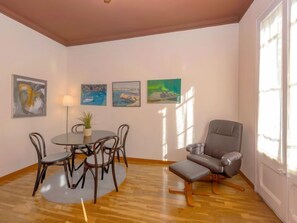 Une pièce comprenant une table à manger ronde, quatre chaises, un fauteuil gris et un petit pouf. Quatre tableaux ornent le mur, ainsi qu’une lampe et une fenêtre qui laisse entrer la lumière naturelle.