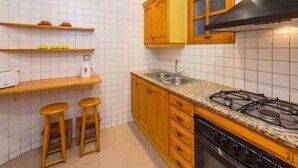 Een keuken met witte betegelde wanden, houten kastjes, een gasfornuis en twee krukjes.