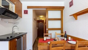 A compact kitchen with a stove, sink, and wooden cabinets. A dining table with chairs is set up in the center of the room. - Chalet Club (Val Claret) by Interhome (Tignes)