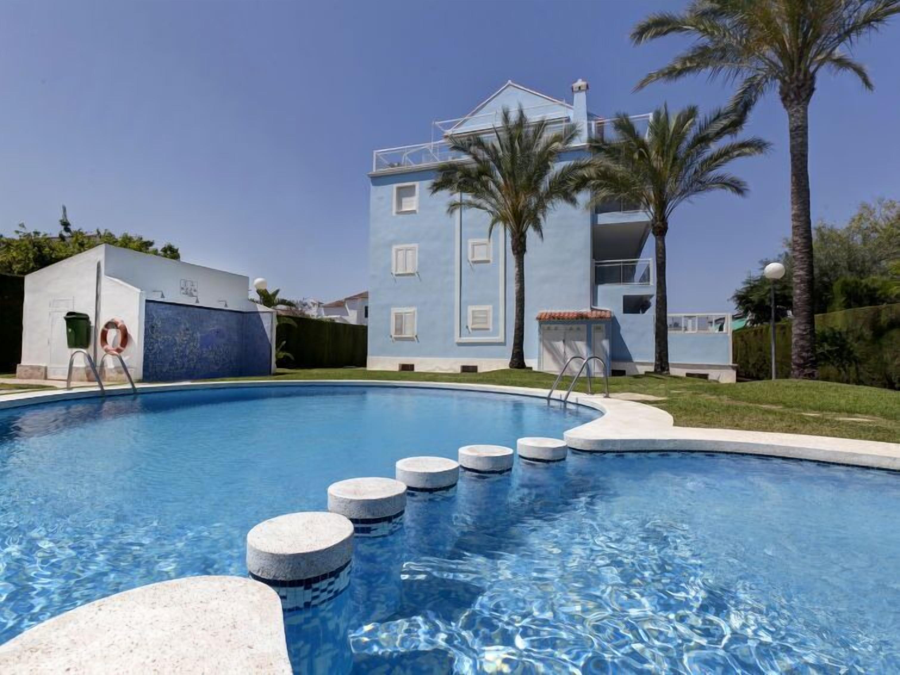 A swimming pool with a clear blue water surface, surrounded by a white building with a blue facade and a balcony.