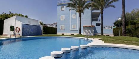 A swimming pool with a clear blue water surface, surrounded by a white building with a blue facade and a balcony.