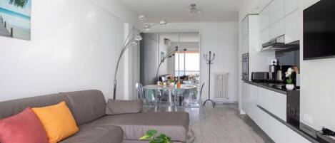 A modern living room with a grey sofa, a small white table, and a potted plant. A dining area with a glass table and chairs is visible in the background.