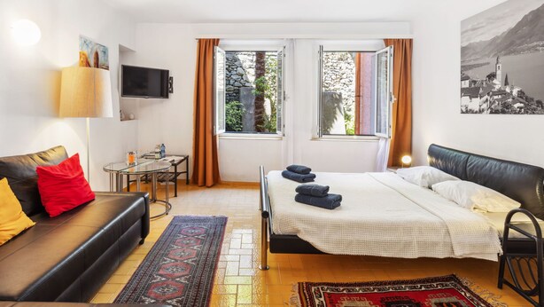 A modern hotel room with a bed, a sofa, a TV, and a window with curtains. - Casa Eduardo Apt.1 by Interhome (Ronco sopra Ascona)